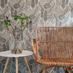 An end table with flower and a woven chair stand in front of a wall papered in a large-scale black and white floral print.
