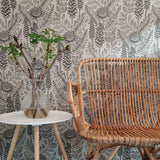 An end table with flower and a woven chair stand in front of a wall papered in a large-scale black and white floral print.