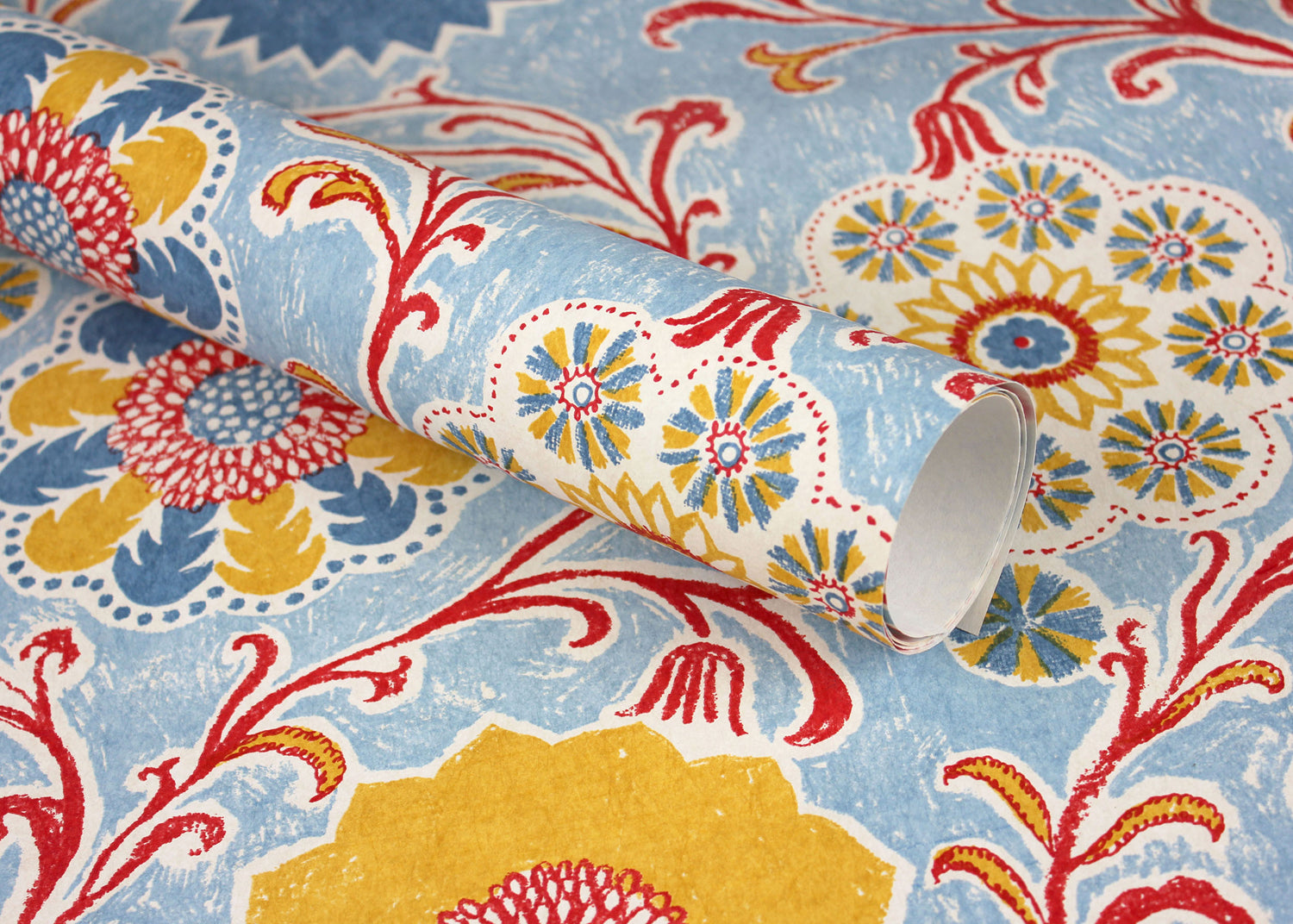 Wallpaper roll in a handdrawn playful floral design in red, yellow, green and blue on a light blue field.