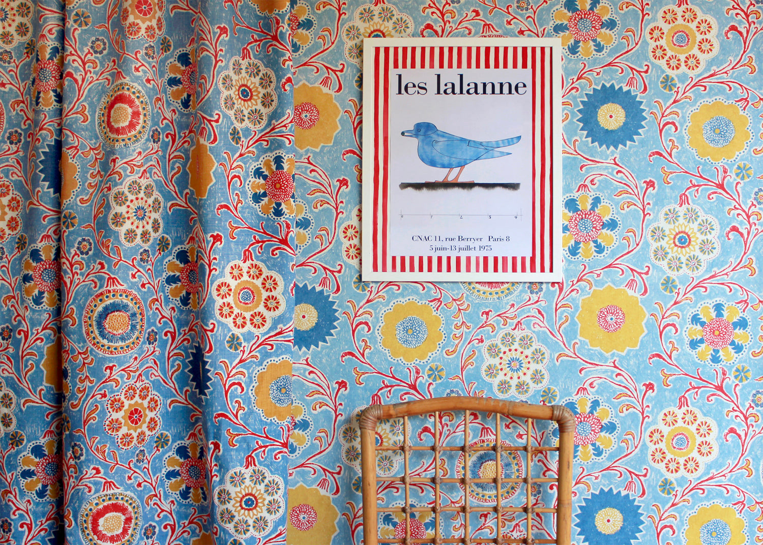 Curtain and wallpaper in matching handdrawn playful floral design in red, yellow, green and blue on a light blue field.