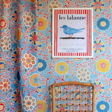 Curtain and wallpaper in matching handdrawn playful floral design in red, yellow, green and blue on a light blue field.