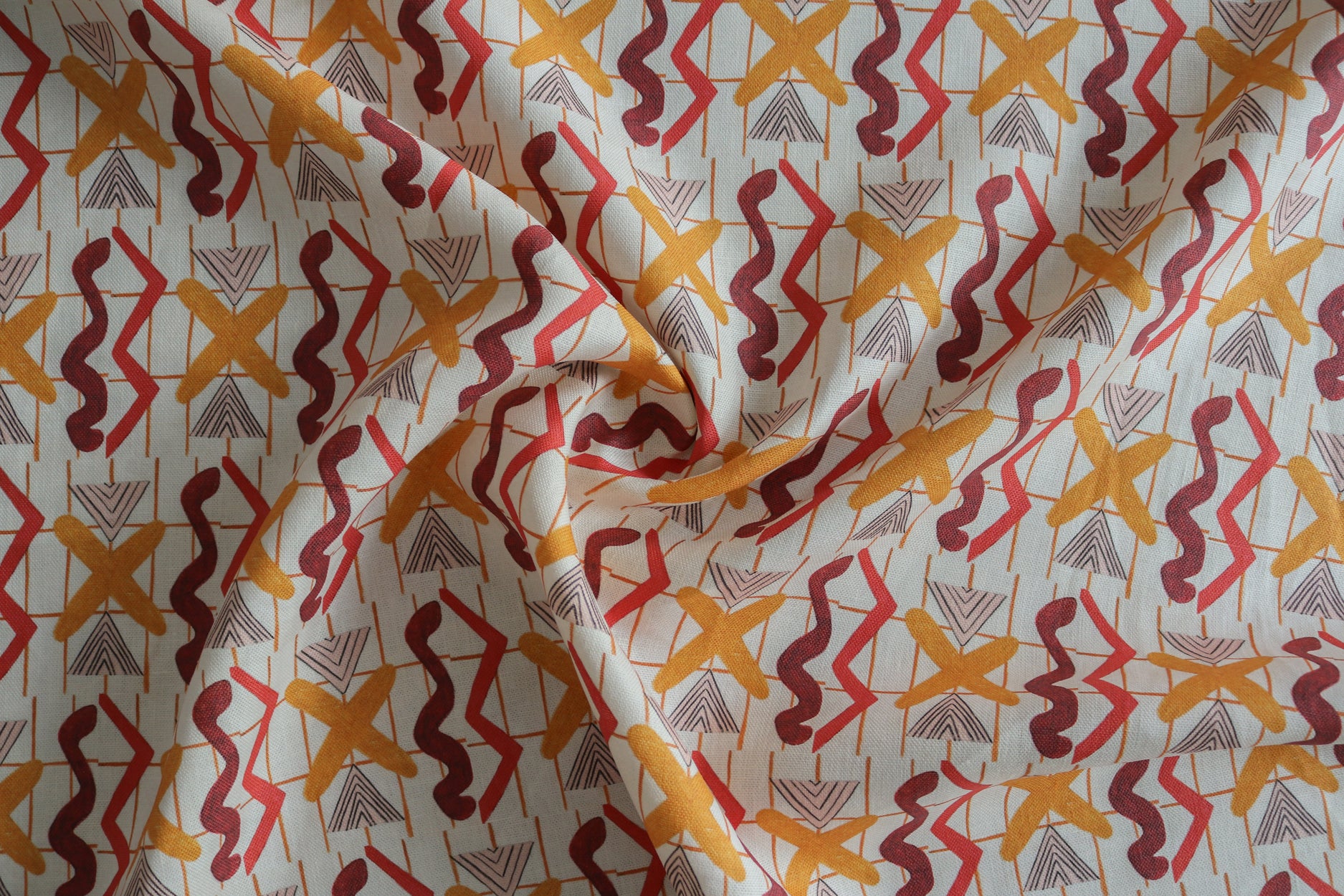 Close up of gently draped printed fabric with red squiggles, pink striped triangles, orange  X marks on a broken grid in orange on a white background. 