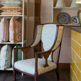 Upholstered wooden side chair in a blue floral against wooden shelving with decorative pillows, handpainted ceramics and lampshades. 