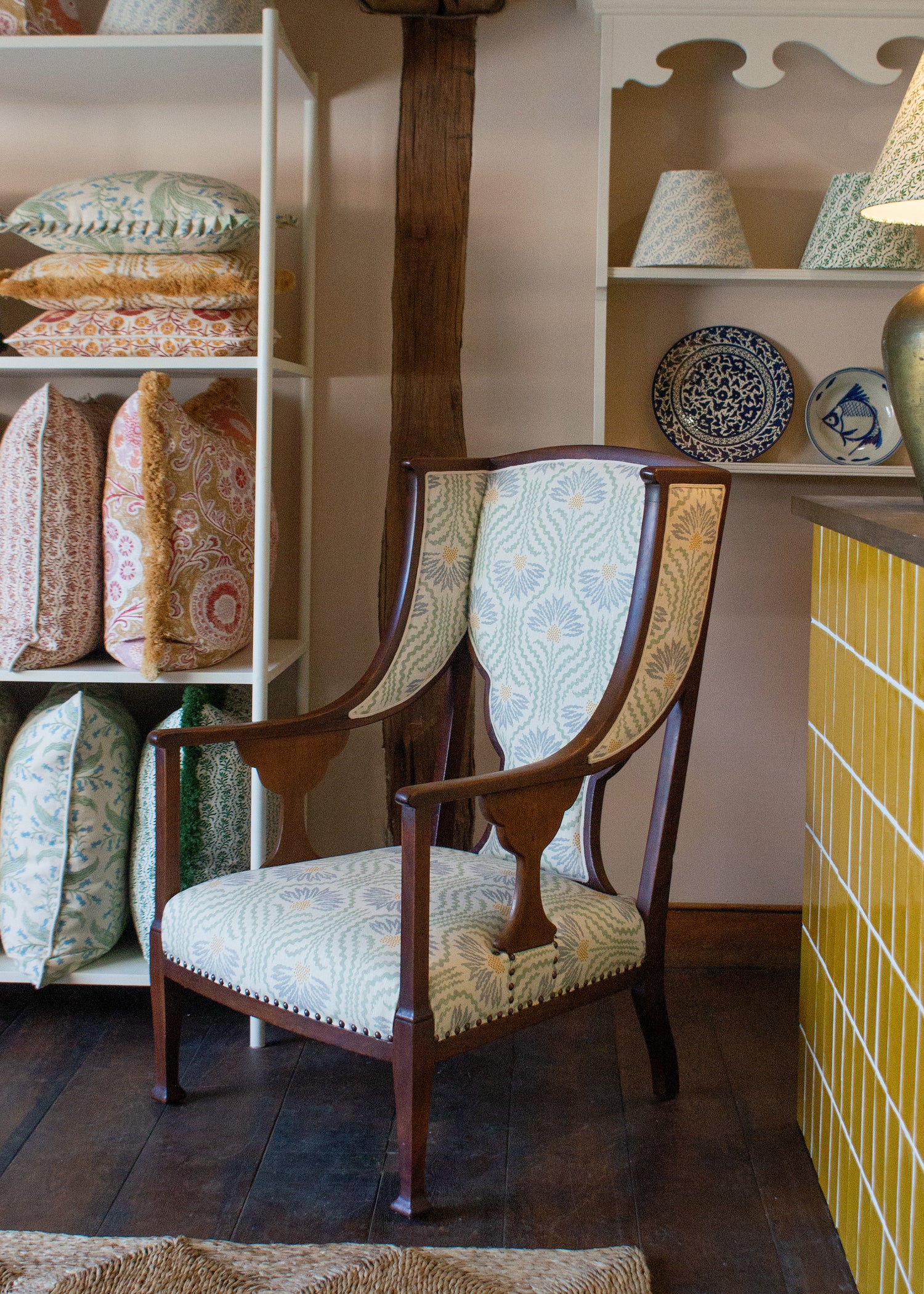 Upholstered wooden side chair in a blue floral against wooden shelving with decorative pillows, handpainted ceramics and lampshades. 