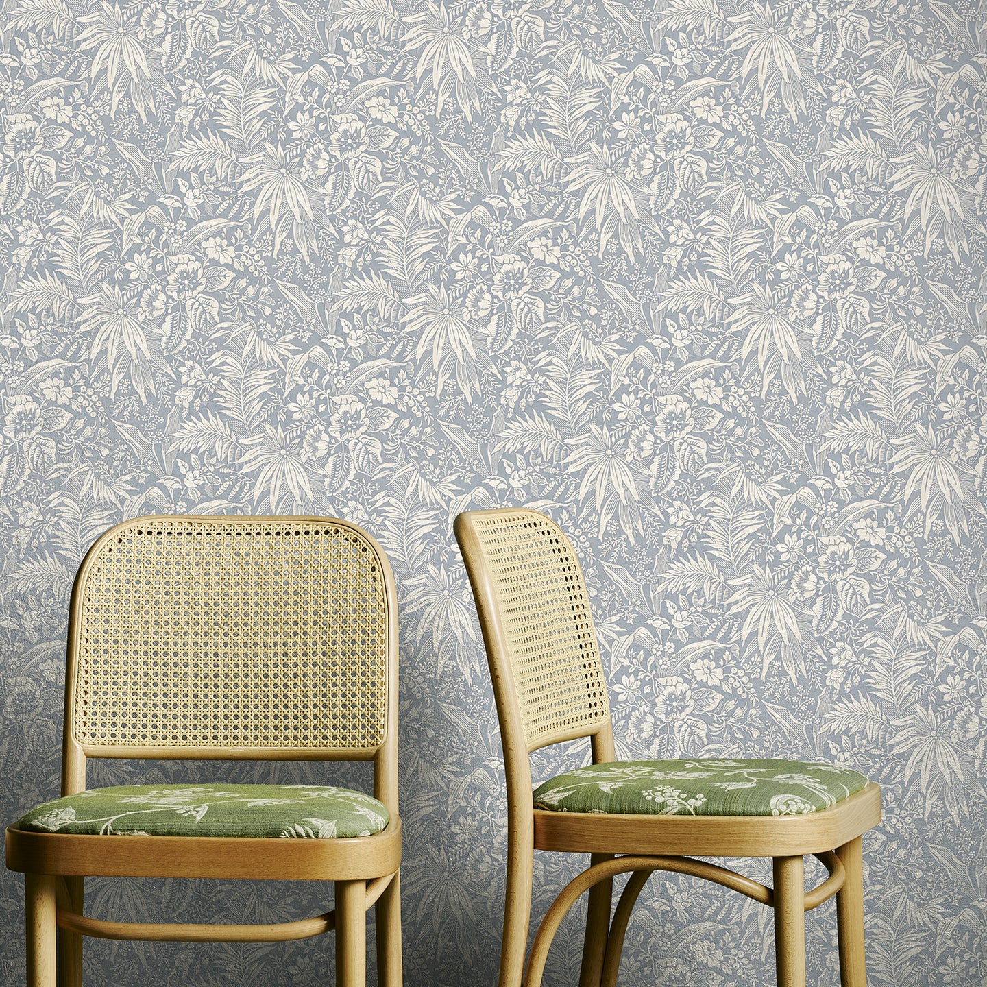 Two wooden chairs with wicker backs against a pale blue floral wallpapered wall.