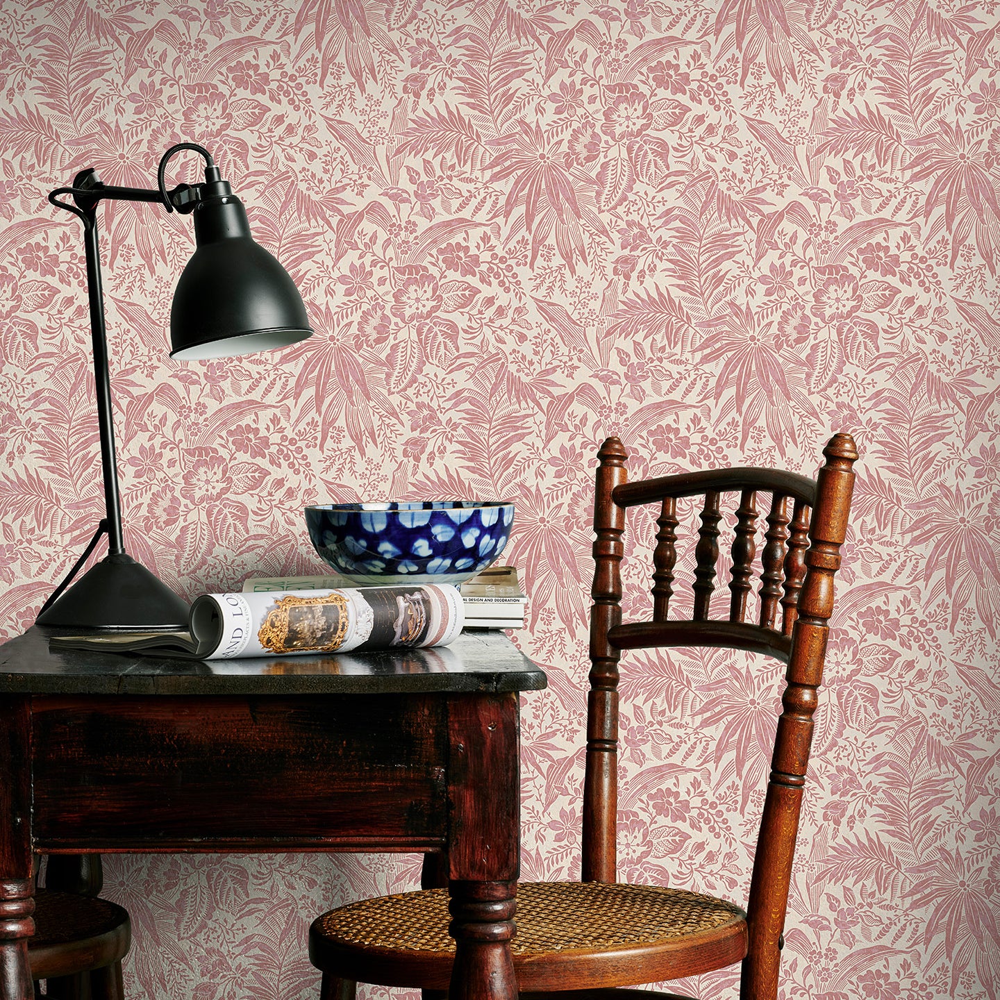Vintage-style room with pink floral wallpaper, wooden chair, and table with lamp and newspaper.