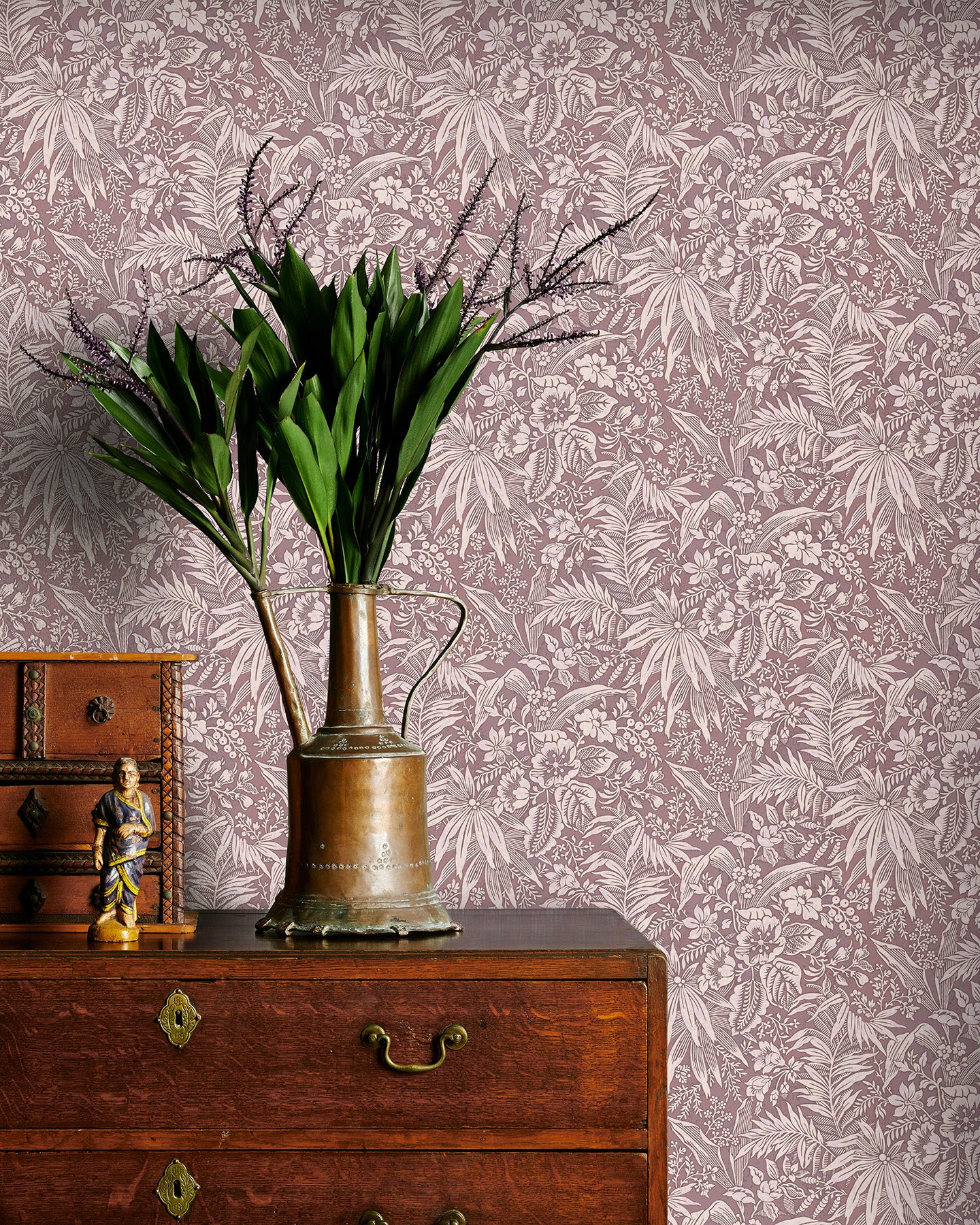 Vintage brass watering can with plants on a wooden dresser against a mauve floral wallpapered wall.