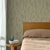 Bed with tan linens and wooden headboard, wooden side table  with white book and green glass, white floor length curtain against a black pointillist wheat frond patterned wallpaper on a sage green background. 