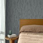 Bed with tan linens and wooden headboard, wooden side table  with white book and green glass, white floor length curtain against a black pointillist wheat frond patterned wallpaper on a steel blue-grey background. 