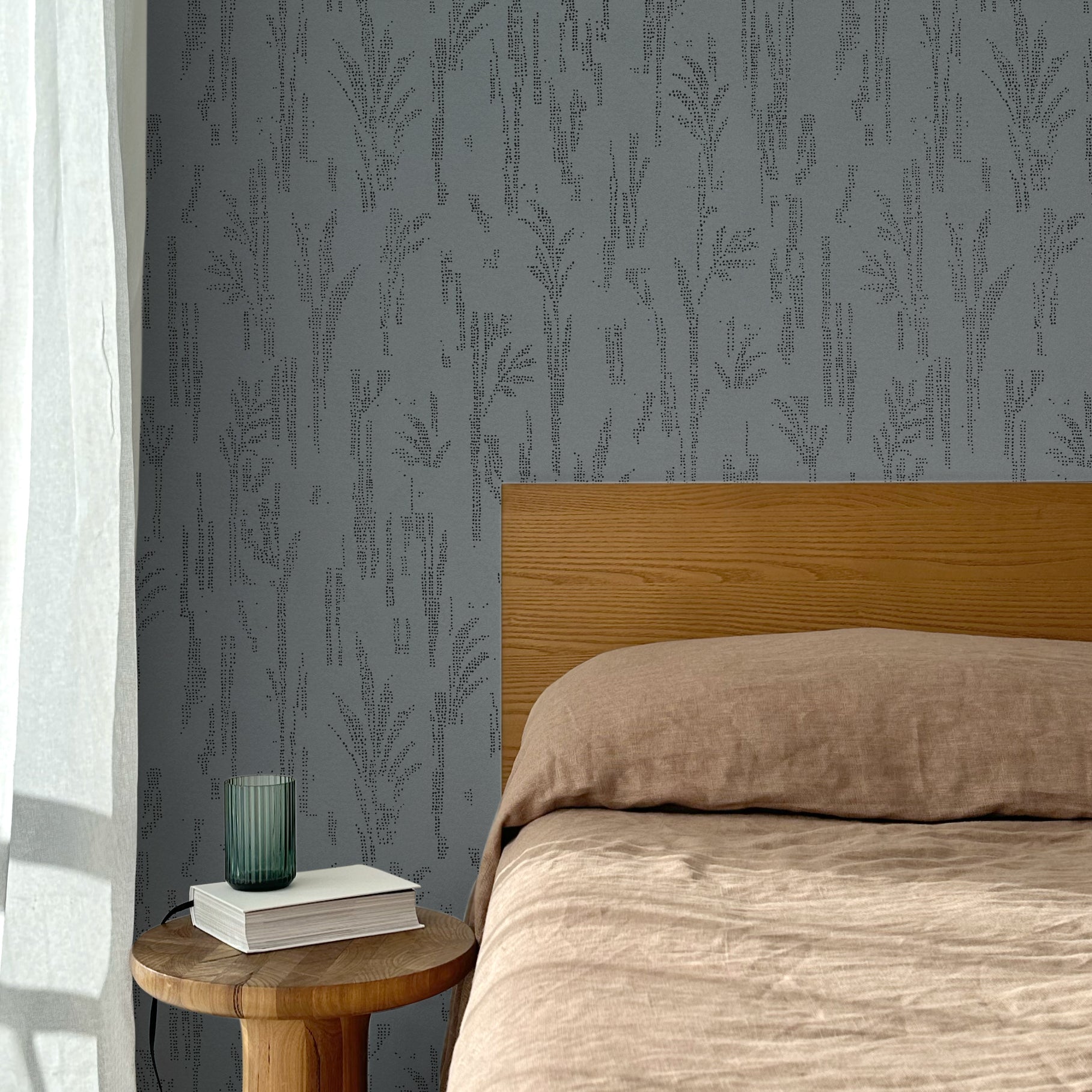 Bed with tan linens and wooden headboard, wooden side table  with white book and green glass, white floor length curtain against a black pointillist wheat frond patterned wallpaper on a steel blue-grey background. 