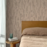 Bed with tan linens and wooden headboard, wooden side table  with white book and green glass, white floor length curtain against a black pointillist wheat frond patterned wallpaper on a soft clay pink background.