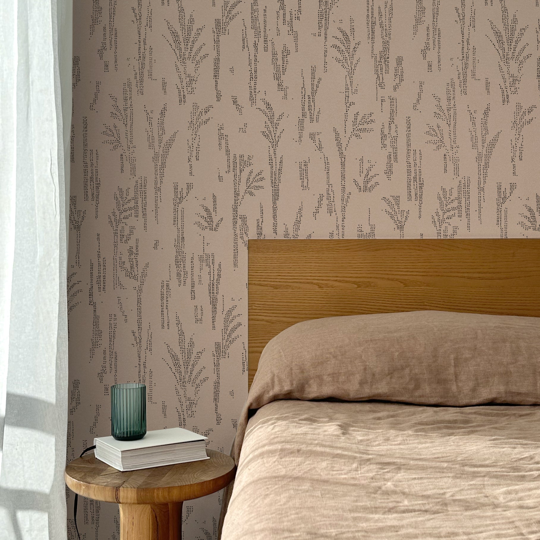 Bed with tan linens and wooden headboard, wooden side table  with white book and green glass, white floor length curtain against a black pointillist wheat frond patterned wallpaper on a soft clay pink background.