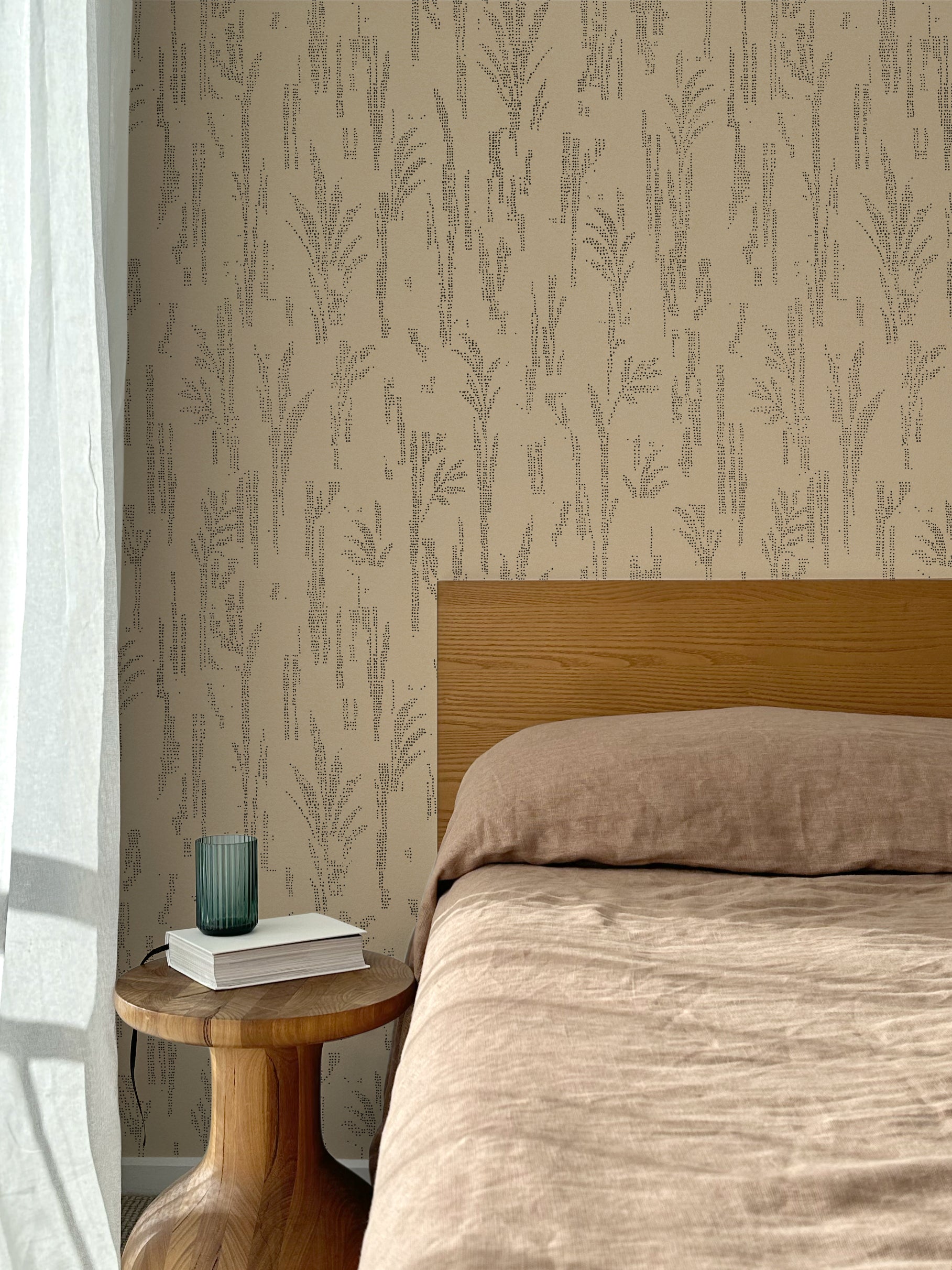 Bed with tan linens and wooden headboard, wooden side table  with white book and green glass, white floor length curtain against a black pointillist wheat frond patterned wallpaper on a tan background. 