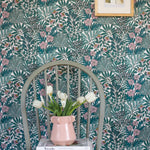 A wooden chair stands in front of a wall papered in a dense floral print in turquoise, pink and yellow.