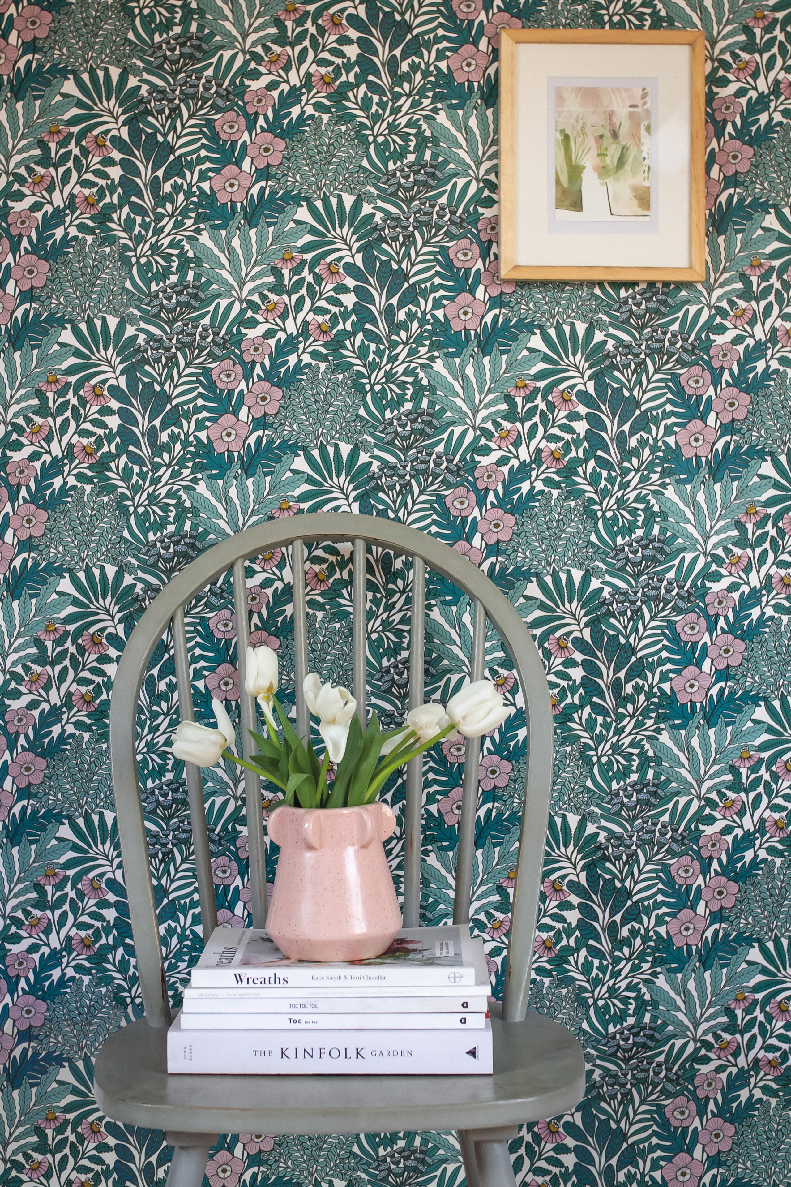 A wooden chair stands in front of a wall papered in a dense floral print in turquoise, pink and yellow.