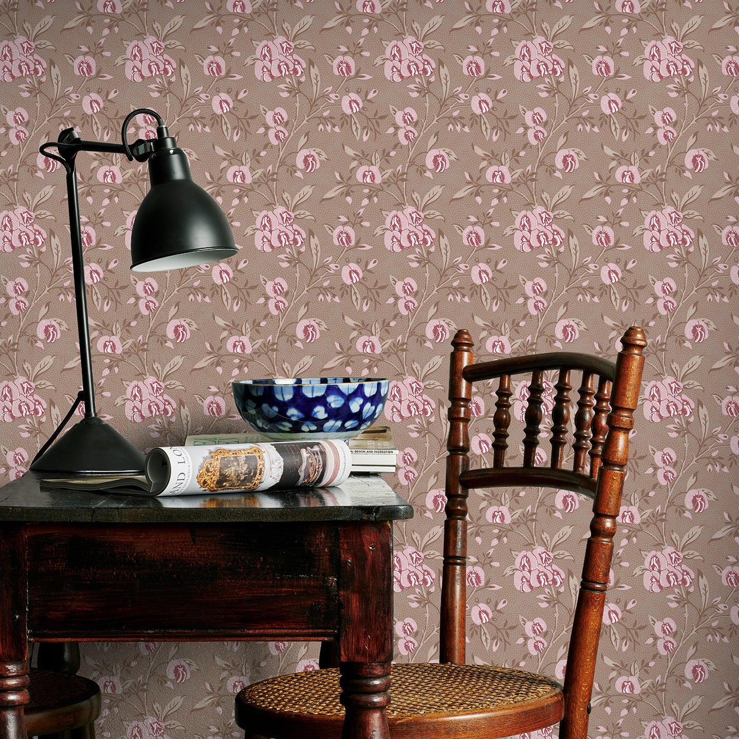 Vintage-style room with brown and pink floral wallpaper, wooden chair, and table with lamp and books.