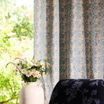 Decorative curtain with floral pattern next to a chair and vase with flowers by a window.