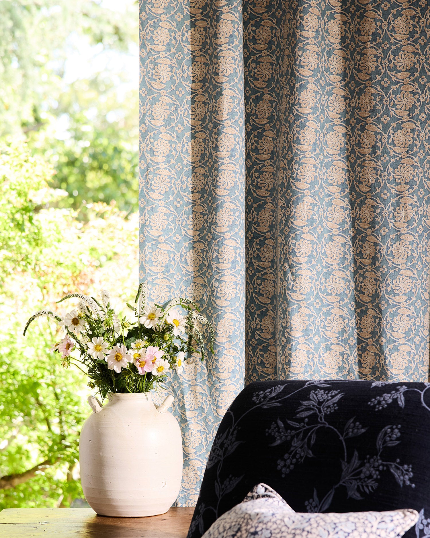 Decorative curtain with floral pattern next to a chair and vase with flowers by a window.