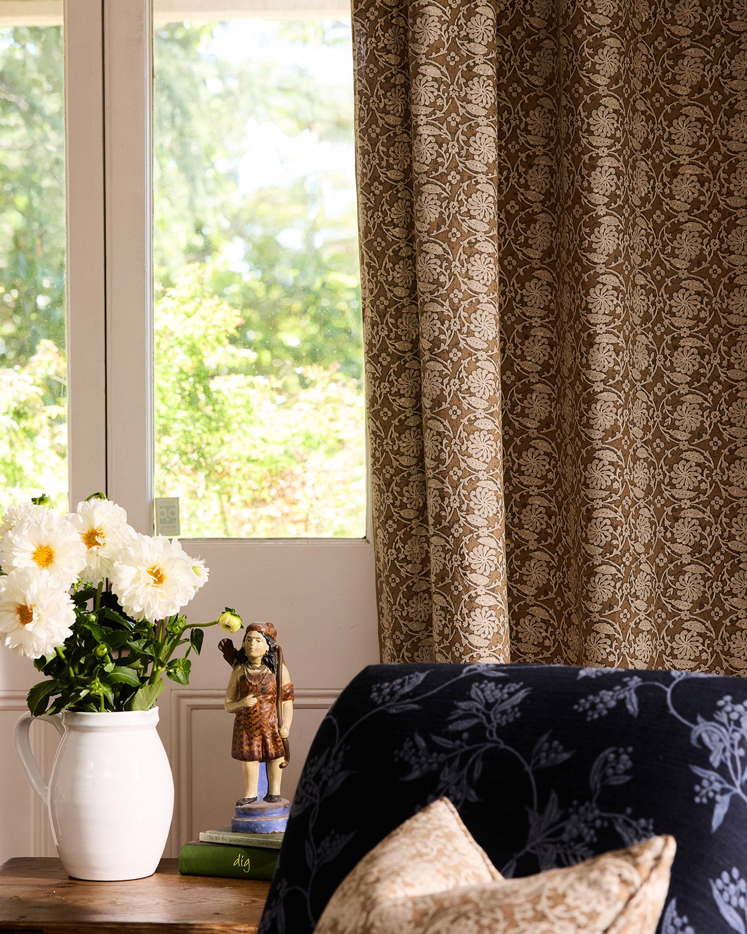 Decorative curtain with floral pattern in a room with a window and a vase of flowers on a table.