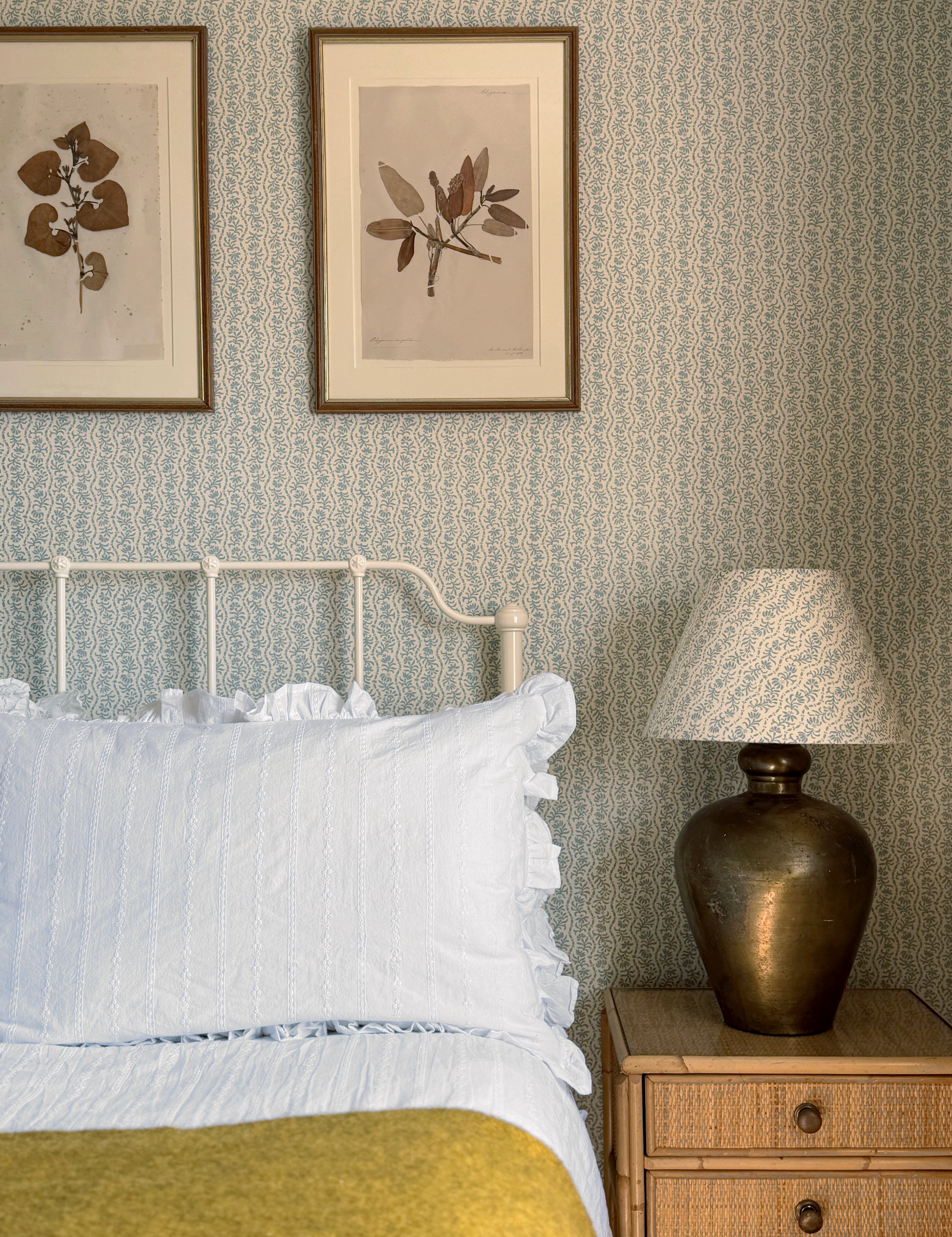 A styled bedroom corner featuring a white metal bed frame with white bedding and a mustard yellow throw, set against a light blue patterned wallpaper. A wooden nightstand with a bronze lamp sits on the right, and two framed botanical prints hang on the wall above the headboard.