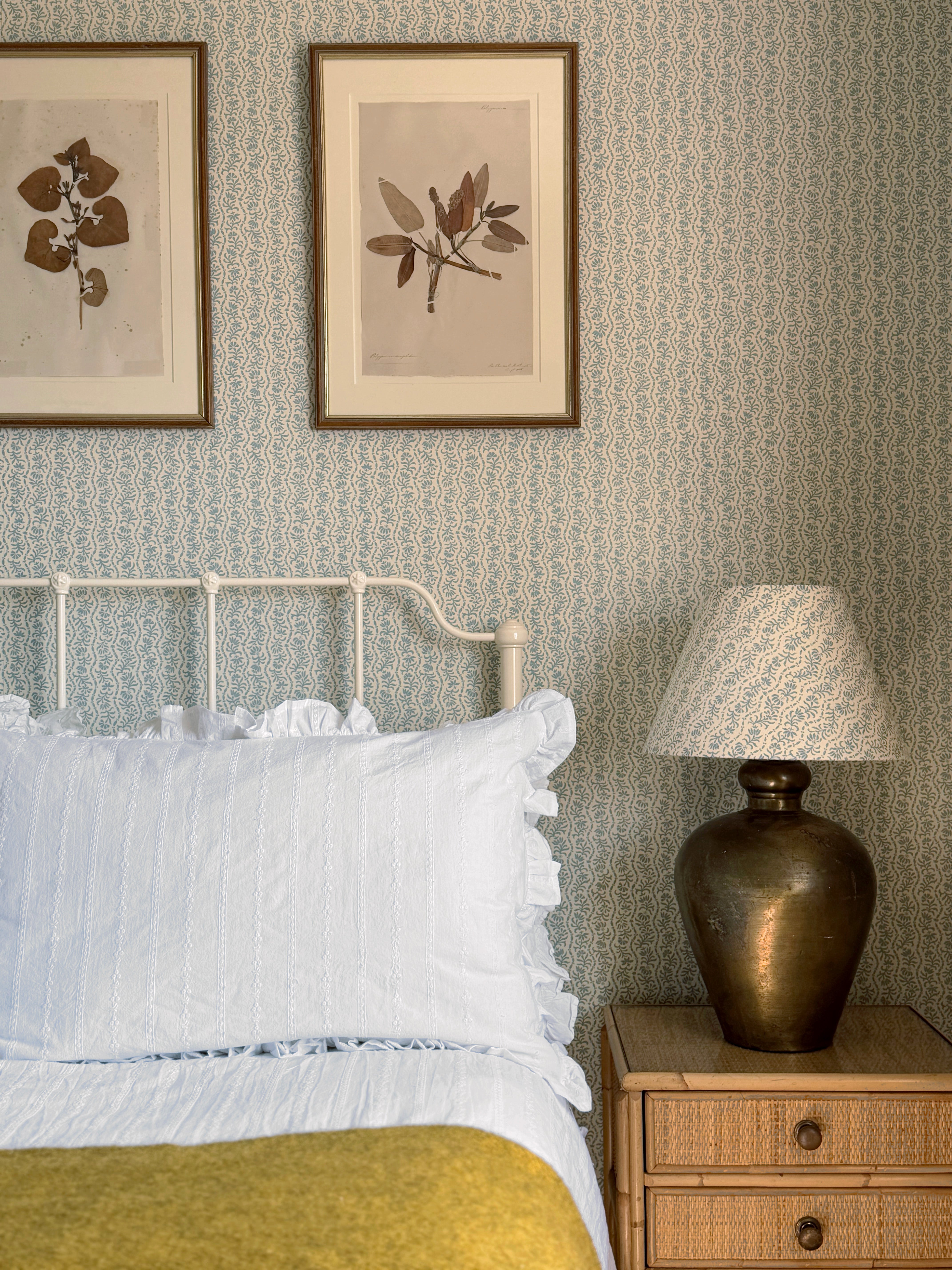 A styled bedroom corner featuring a white metal bed frame with white bedding and a mustard yellow throw, set against a light blue patterned wallpaper. A wooden nightstand with a bronze lamp sits on the right, and two framed botanical prints hang on the wall above the headboard.