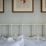 Light blue and white floral stripe patterned wallpaper on a bedroom wall above a white metal bed frame with white ruffled bedding. Three framed botanical prints hang on the wall.