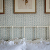 Light blue and white floral stripe patterned wallpaper on a bedroom wall above a white metal bed frame with white ruffled bedding. Three framed botanical prints hang on the wall.
