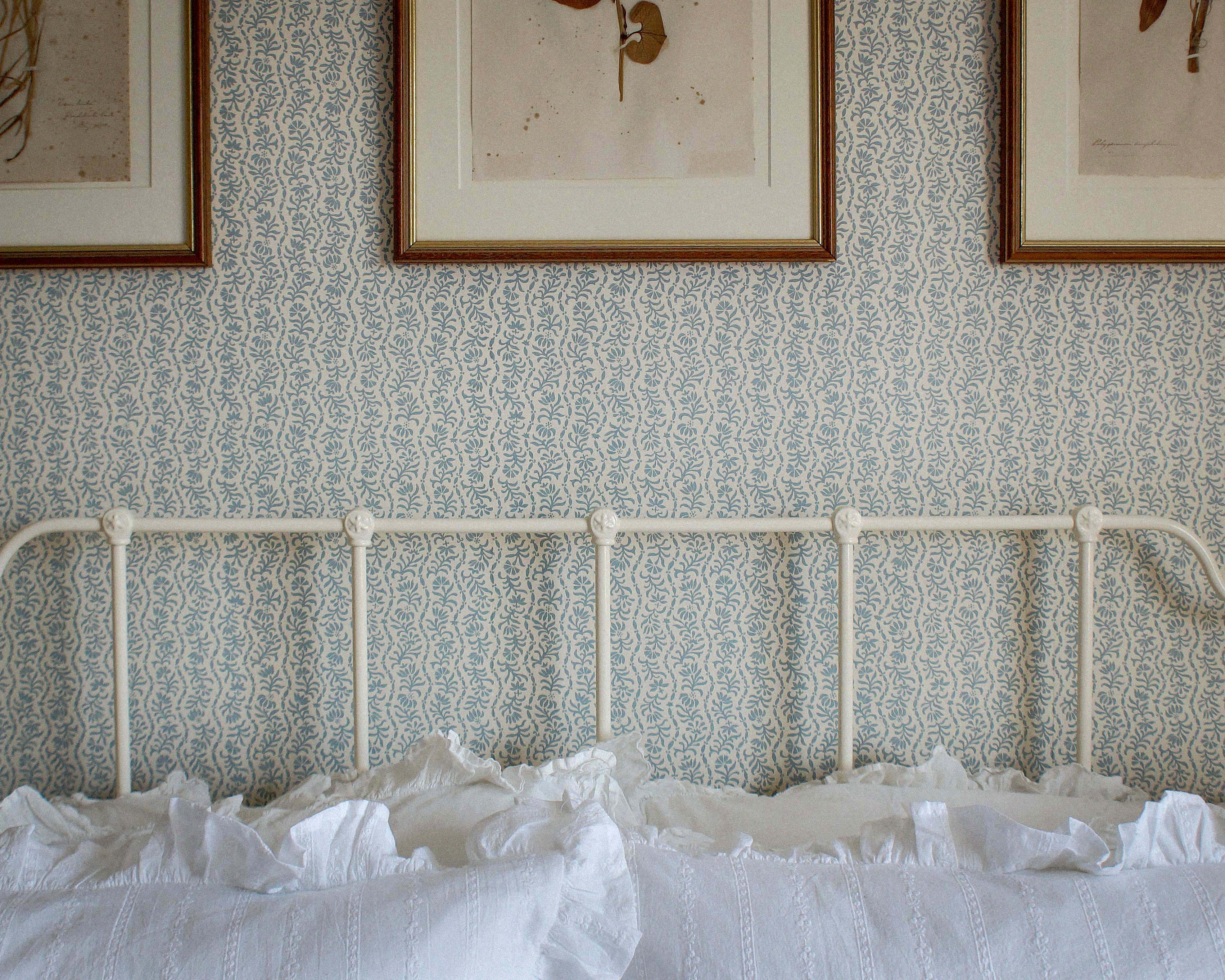 Light blue and white floral stripe patterned wallpaper on a bedroom wall above a white metal bed frame with white ruffled bedding. Three framed botanical prints hang on the wall.