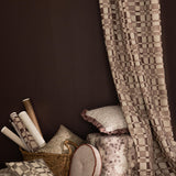 Detail of a styled interior scene with a floor to ceiling draped fabric with a brown and white checkerboard along side stacked throw pillows, ottoman and basket with wallpaper rolls in various patterns in a range of muted colors against a dark brown wall. 