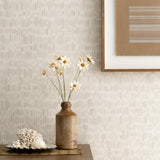 Detail of a styled interior scene set against wallpaper with a soft beige a watercolor broken stripe pattern featuring a wooden dresser, ceramic vase with daisies, dish with seashells against a wall with a framed brown and white abstract line drawing. 