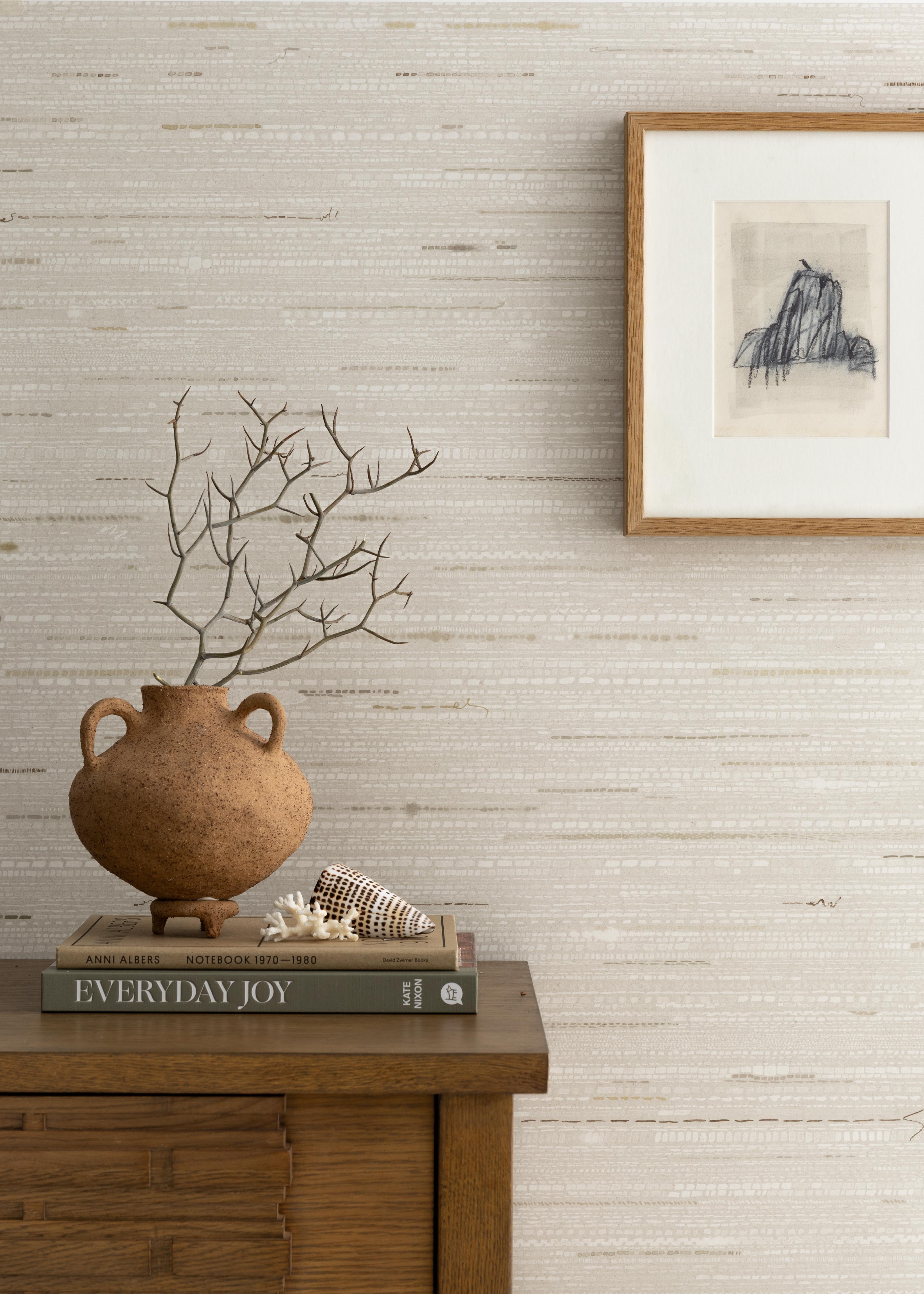 Detail of a styled interior scene set against wallpaper in a grey dashed stripe with a framed abstract charcoal drawing, wooden dresser with two hardcover books and a ceramic urn with handles and a dried branch, along side a piece of white coral and large seashell.