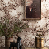Detail of a styled interior scene set against wallpaper in a brown watercolor leaf pattern with a framed abstract painting featuring a wooden table with a ceramic vase with greenery and a black ceramic horse sculpture and a brass canteen and rattan placemat. 