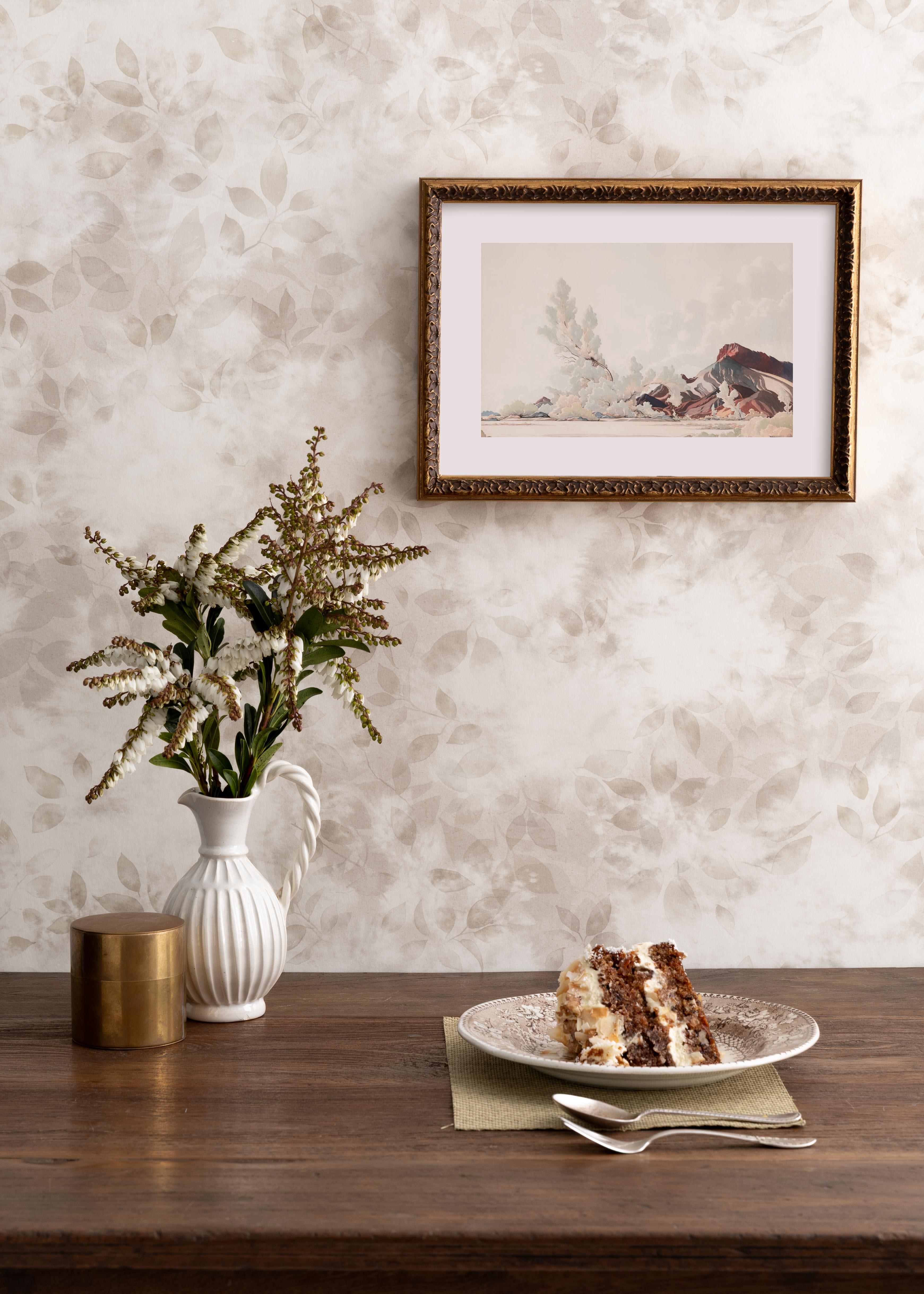 Detail of a styled interior scene set against wallpaper with a beige watercolor leaf pattern with a framed landscape print behind a wooden table with ceramic pitcher with lilies of the valley and brass canister, and a ceramic plate with a slice of cate on a napkin with two spoons. 