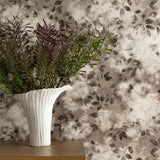Detail of a styled interior scene set against wallpaper with a warm charcoal grey watercolor leaf pattern behind a wooden dresser with a white ceramic vase with greenery. 