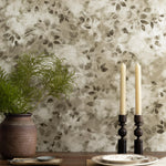 Detail of a styled interior scene set against wallpaper with a muted green watercolor leaf pattern behind a wooden table with ceramic vase with greenery, ornamental spoon, ornate candle sticks and a single place setting with stacked plates and a draped napkin. 