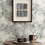 Detail of a styled interior scene set against wallpaper with a grey watercolor leaf pattern featuring a framed stacked abstract prints and an antique wooden table with ceramic mug and plate with pastries and a serving spoon. 