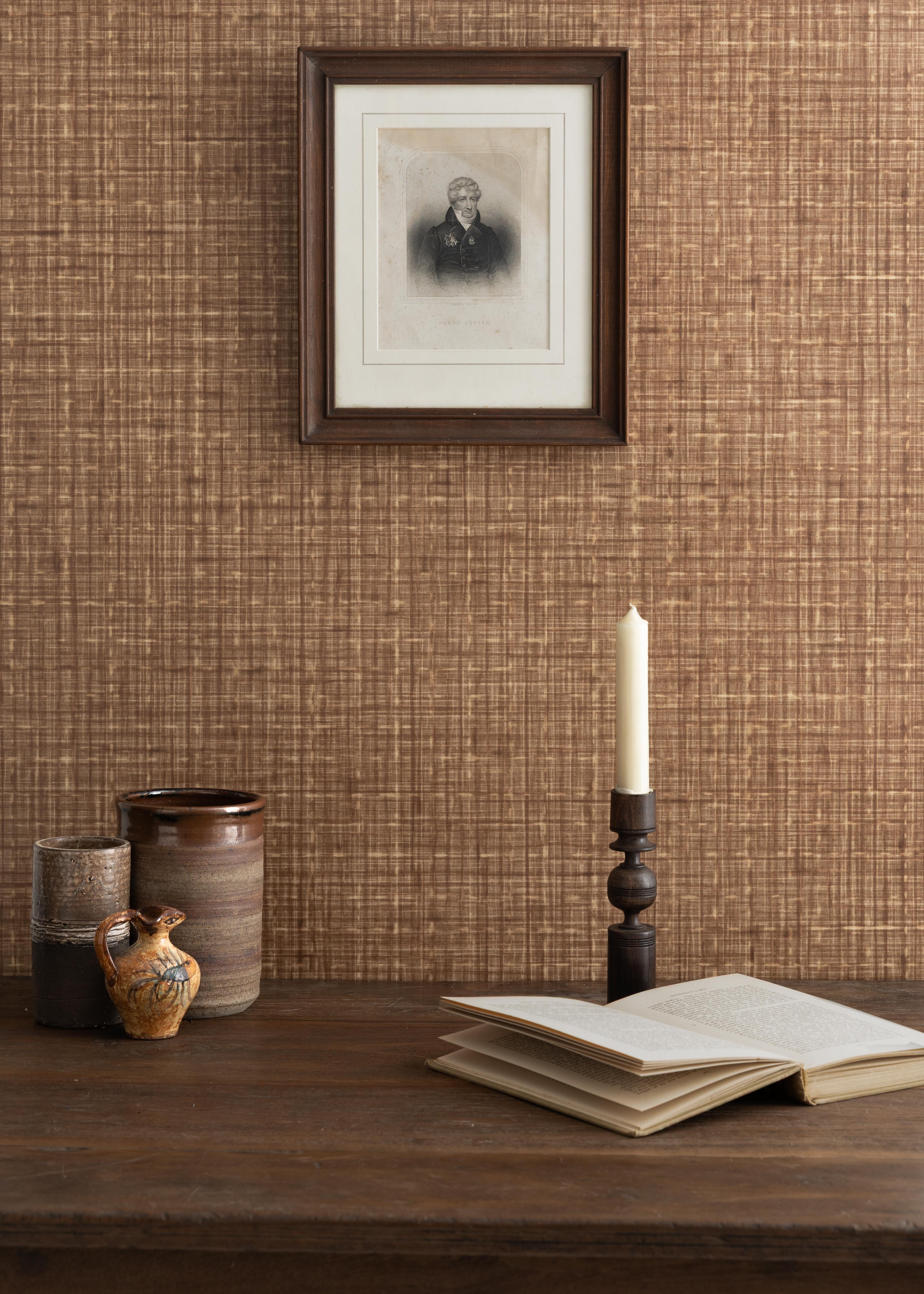 Detail of a styled tabletop scene with ceramic cups, open book and candlestick against a brown textural wallpaper with framed antique black and white portrait of a man in black and white. 