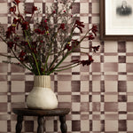 Detail of a styled interior scene set against wallpaper with a wine purple checkerboard design featuring a framed antique portrait of a soldier and a dark wood stool with carved legs and a ceramic vase with red flowers and dried branches.