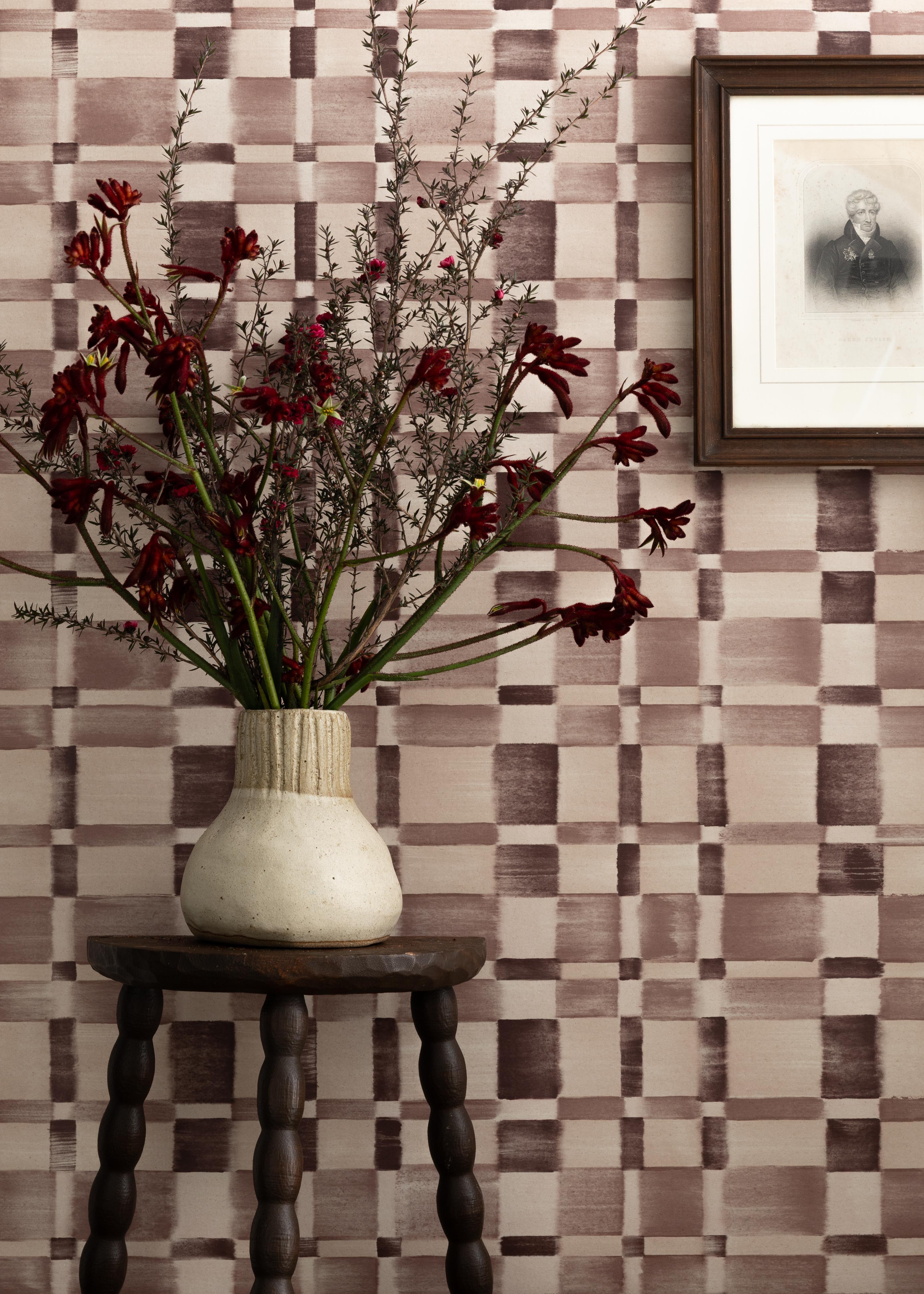 Detail of a styled interior scene set against wallpaper with a wine purple checkerboard design featuring a framed antique portrait of a soldier and a dark wood stool with carved legs and a ceramic vase with red flowers and dried branches.