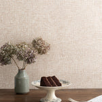 Detail of a styled interior scene set against wallpaper in a watercolor blush pink check with a wooden table featuring dried flowers in a ceramic vase and a white painted cake stand with slide of chocolate cake with fork and knife over a blue patterned napkin. 