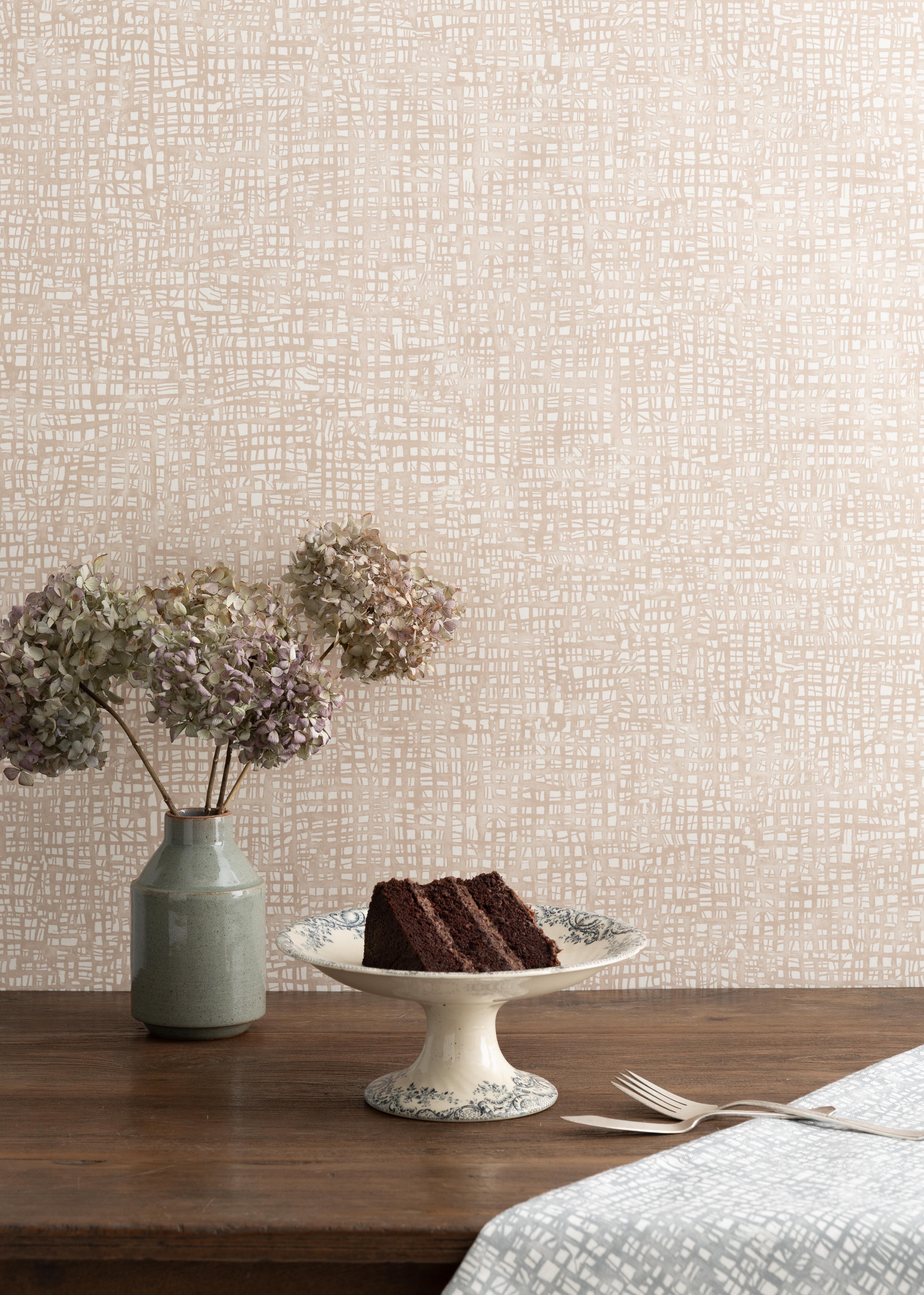 Detail of a styled interior scene set against wallpaper in a watercolor blush pink check with a wooden table featuring dried flowers in a ceramic vase and a white painted cake stand with slide of chocolate cake with fork and knife over a blue patterned napkin. 