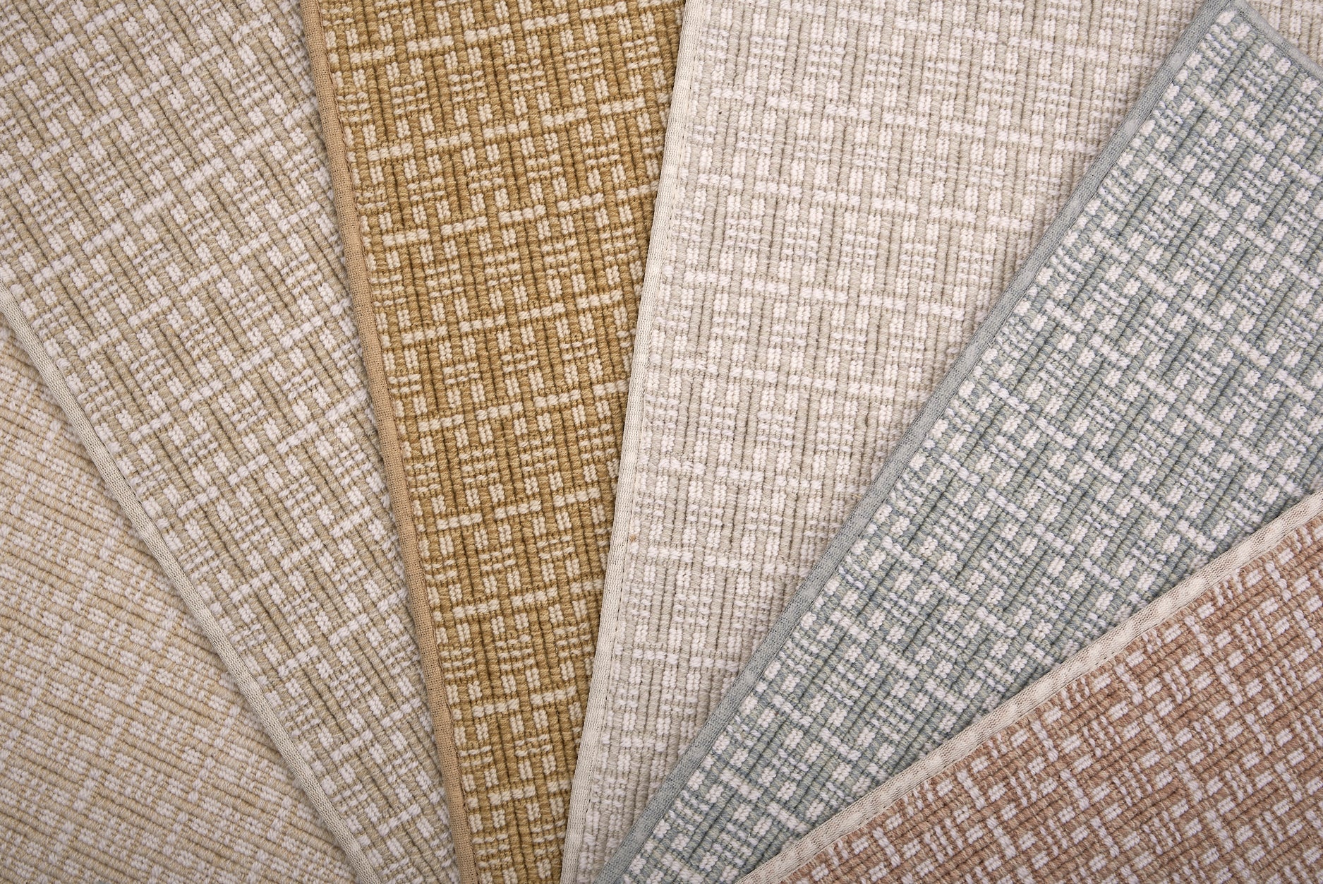 A close-up fan arrangement of basketweave pattern carpet samples in beige, tan, cream, light blue-gray, and dusty rose tones.