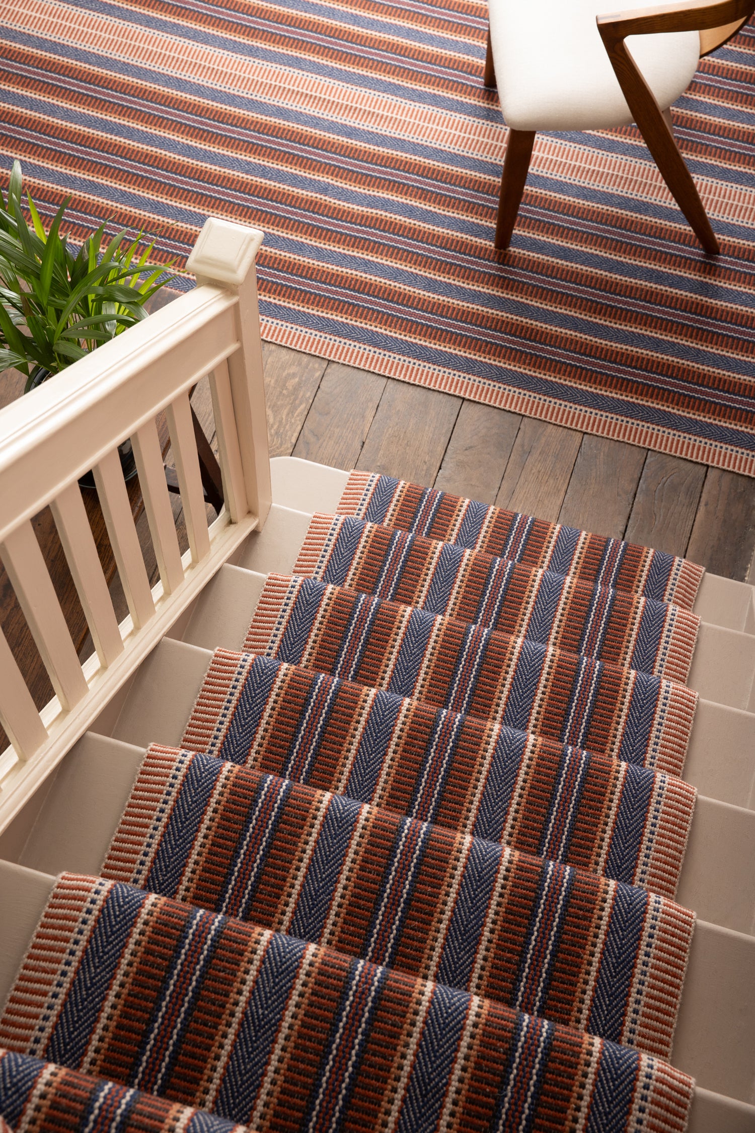 Overhead image of a stair case and landing with a runner and area rug in a complex woven striped made of wool in shades of blue and red. 