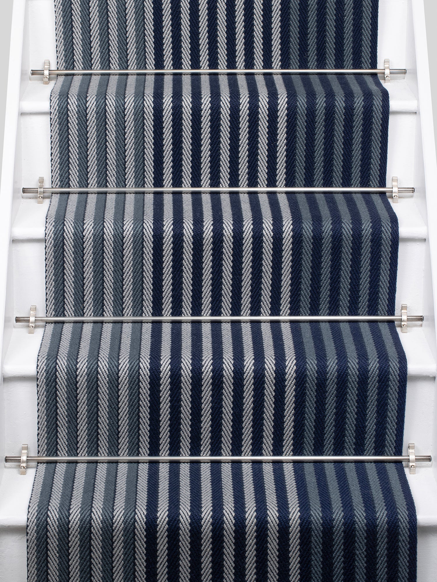 A close-up view of a staircase runner with vertical navy blue, steel and gray herringbone stripes, mounted on white stairs with metal rails.