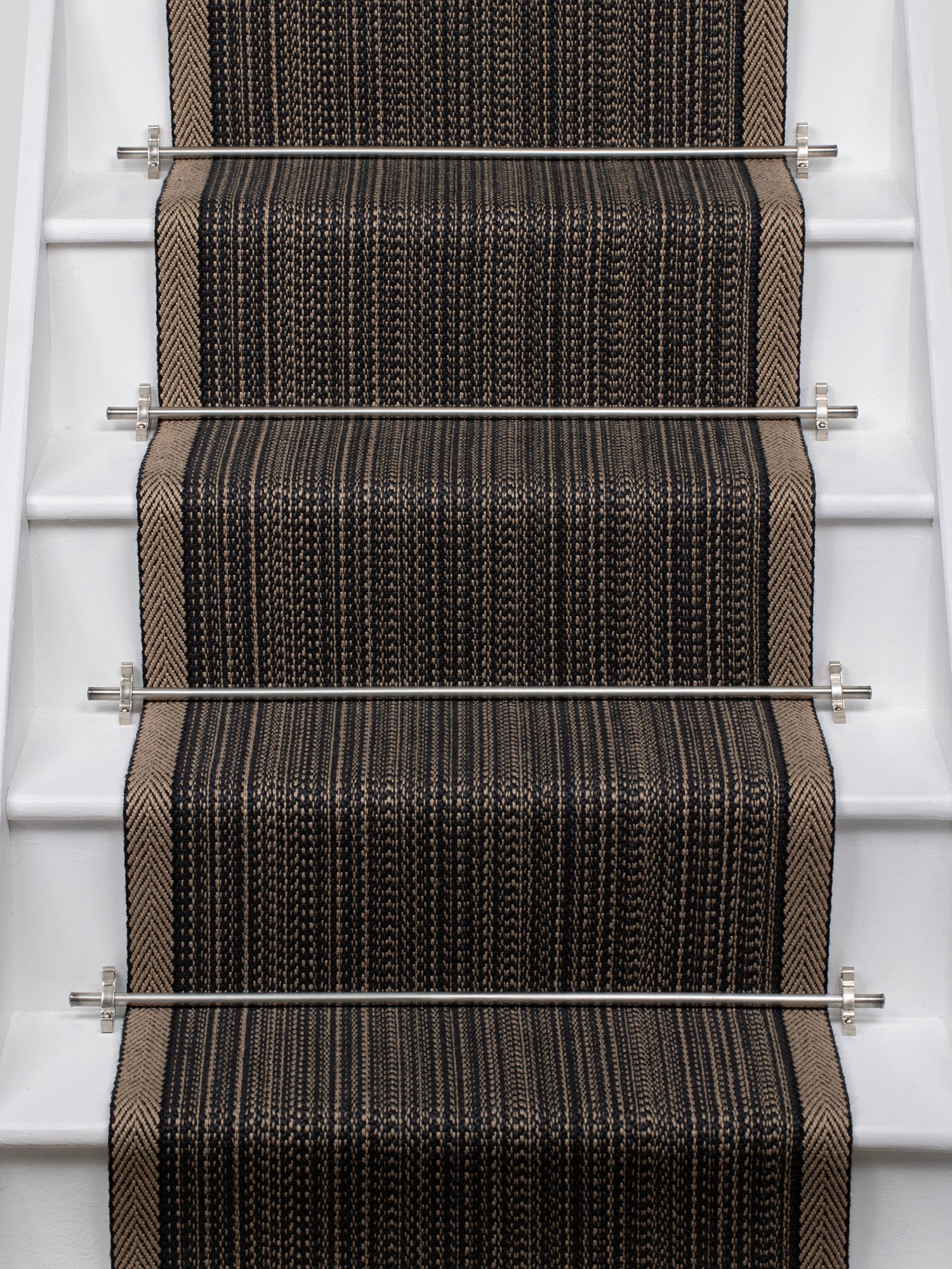 A close-up view of a stair runner carpet installed on a staircase, featuring a textured herringbone pattern in black, brown, and beige with silver metal rails.