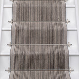 A close-up view of a woven stair runner installed on a staircase, featuring a textured herringbone pattern in gray and beige with stainless steel rails.