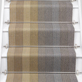 A close-up view of a staircase runner with vertical degradé stripes ranging from beige to yellow to navy blue mounted on white stairs with metal rails.
