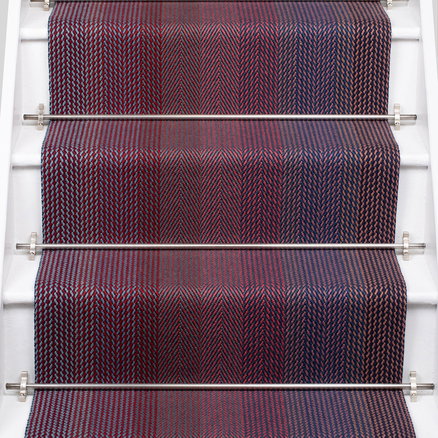 A close-up view of a staircase runner with vertical degradé stripes ranging from deep burgundy, purple, to blue mounted on white stairs with metal rails. 
