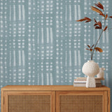 A wooden credenza with a vase and bookes in front of wallpaper in a striped print inspired by Japanese shibori on a soft blue background.