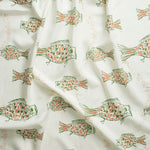 Close up of gently draped printed linen with a pattern of hand-drawn fish in green with tan and orange scales and scattered black bubble dots on white background.